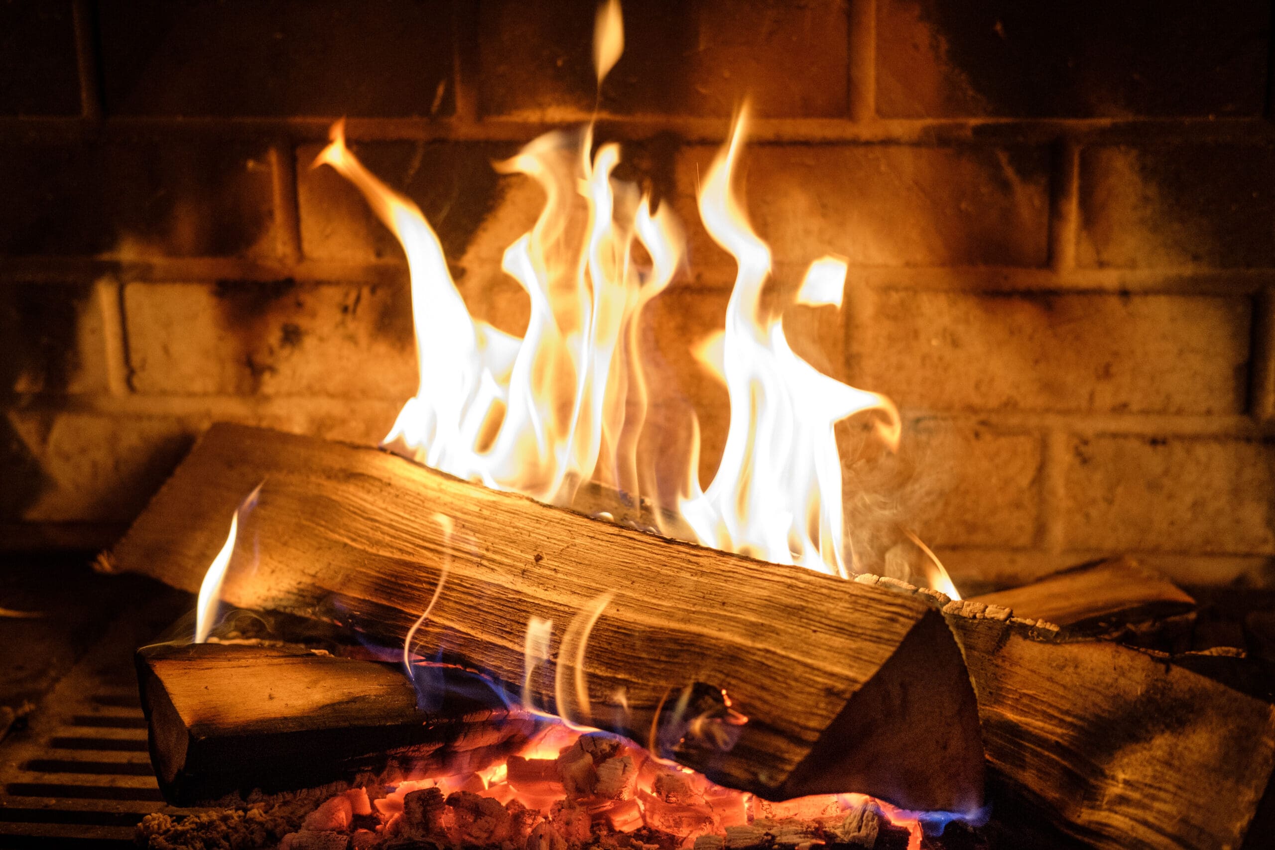 A warm fire burns in a fireplace. Logs are stacked on a metal grate, with bright orange and yellow flames dancing around them. The brick interior of the fireplace glows softly in the background, creating a cozy atmosphere.