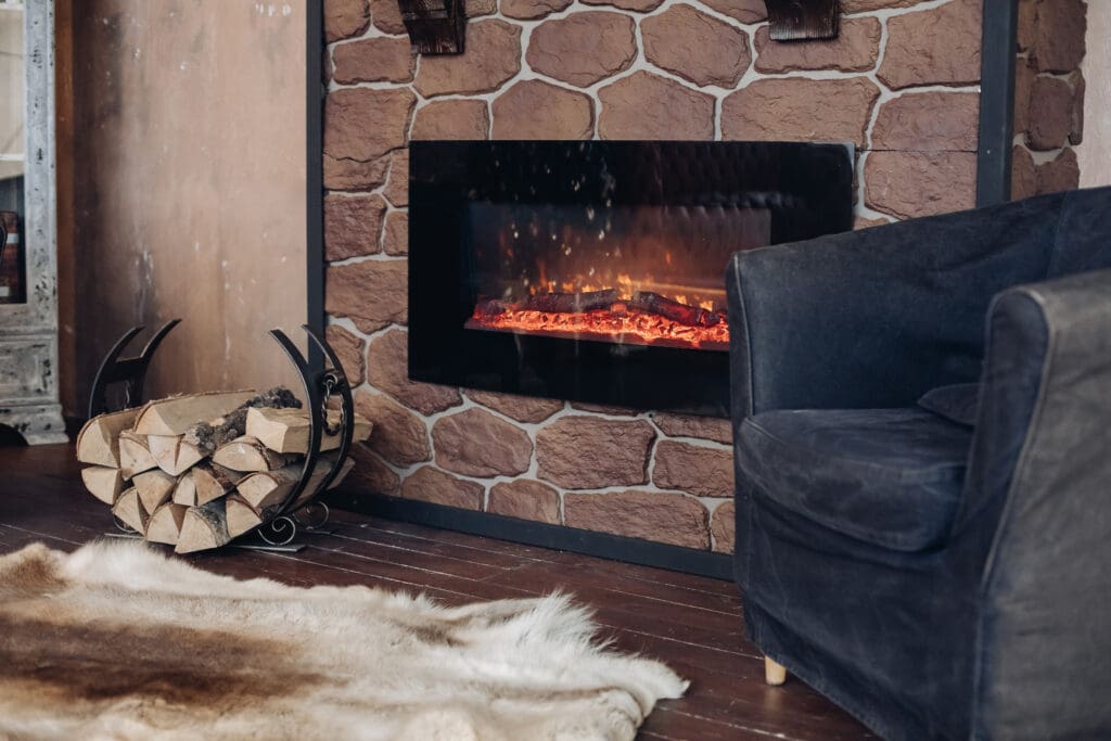 A cozy living room features a modern electric fireplace with a faux stone facade. A black armchair is positioned nearby, and a metal log holder with firewood sits on the wooden floor beside a beige and brown patterned rug.