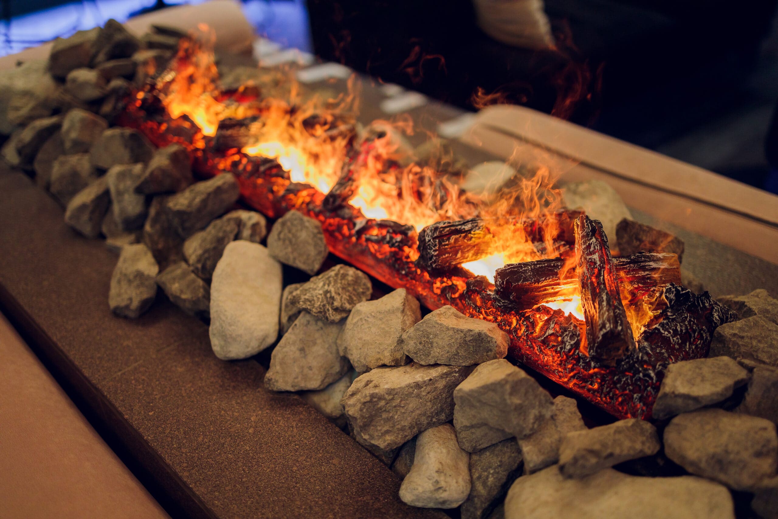 A rectangular tabletop fire pit with realistic-looking flames and logs, surrounded by a neatly arranged border of large, light-colored stones, set on a brown surface.