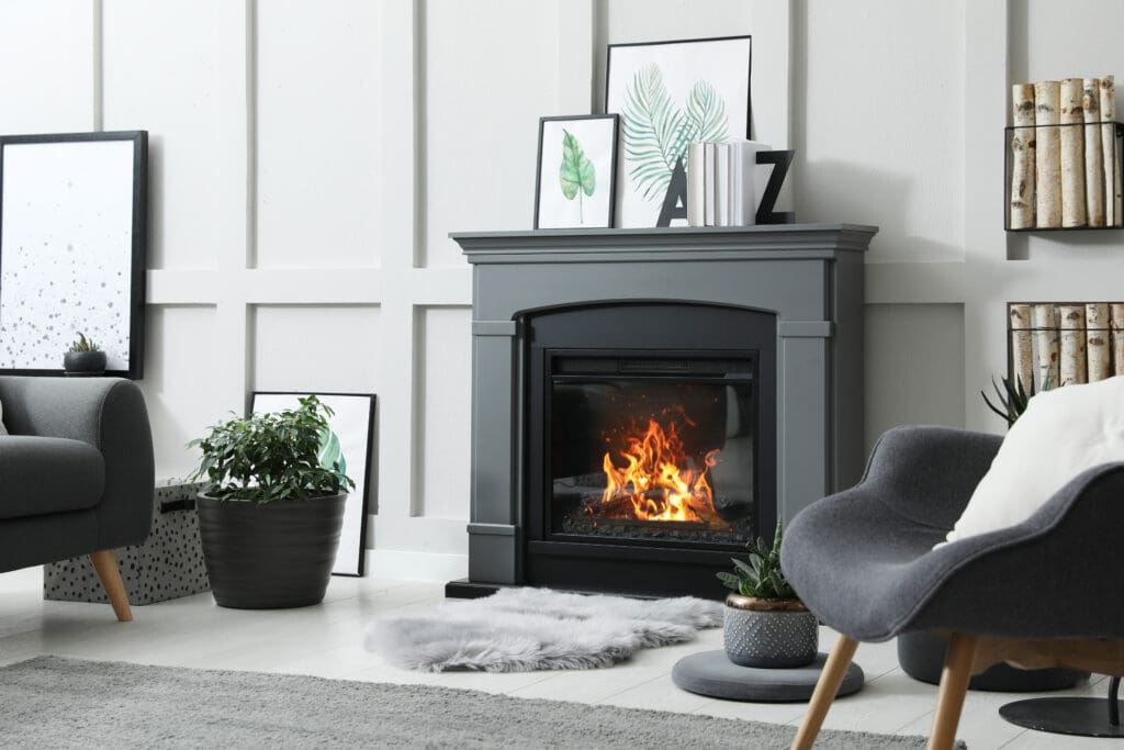 Modern living room with a sleek black fireplace and a burning fire. Grey chair and plush rug in front. Framed art and plants decorate the space, with a stack of firewood beside the mantel. Light-colored walls and floor provide a cozy ambiance.