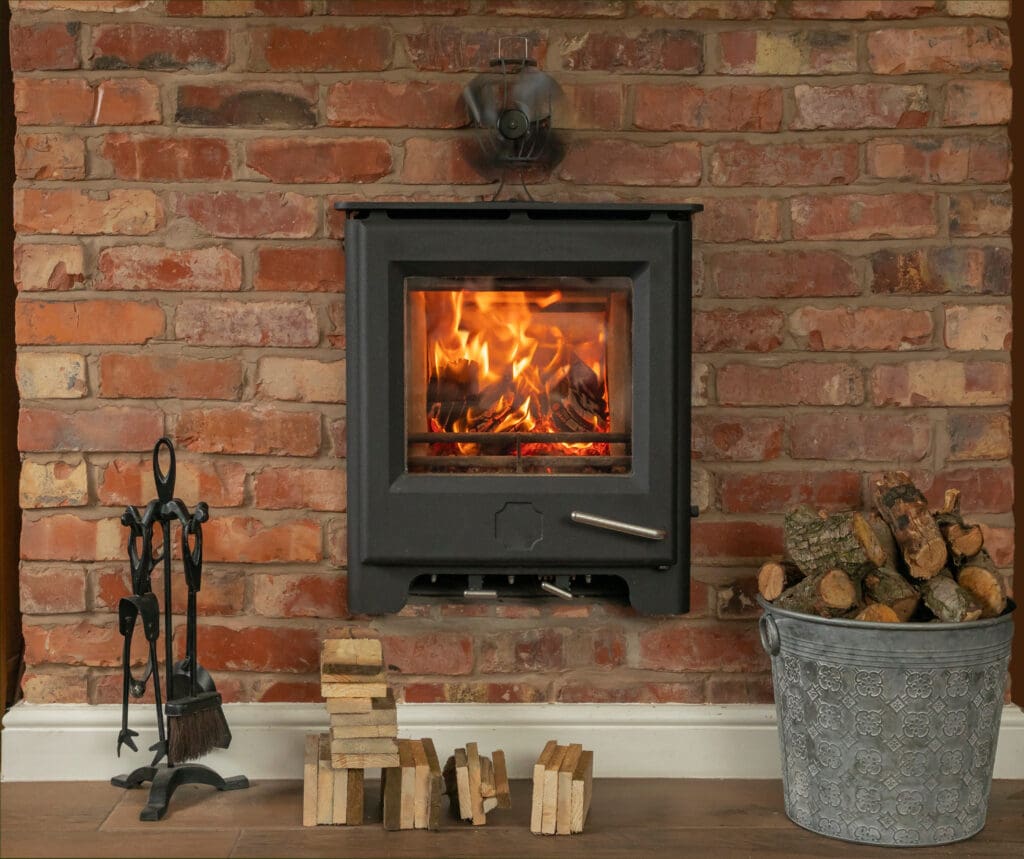 A cozy wood-burning stove set against a rustic brick wall, with a vibrant fire burning inside. Nearby, a metal bucket filled with logs, a stack of cut wood pieces, and a set of fireplace tools add to the warm, inviting atmosphere.