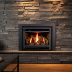 A cozy living room features a modern stone fireplace with a warm fire burning inside. A wooden mantle holds decorative items, and nearby, a table with white ceramics adds to the inviting atmosphere.