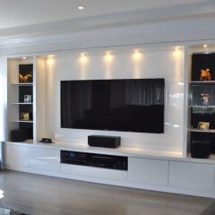 Modern living room with a large flat-screen TV mounted on a white entertainment center. The unit has shelves with books, decorative items, and framed photos. Tall speakers stand on either side. Recessed lighting adds ambiance. Large window on the left.