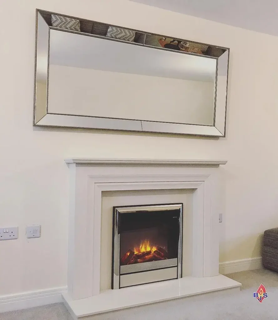 Elegant white fireplace with a modern electric fire insert topped by a large rectangular mirror with a beveled frame. The setup is in a minimalist living room with a light gray wall. Cozy and inviting atmosphere.