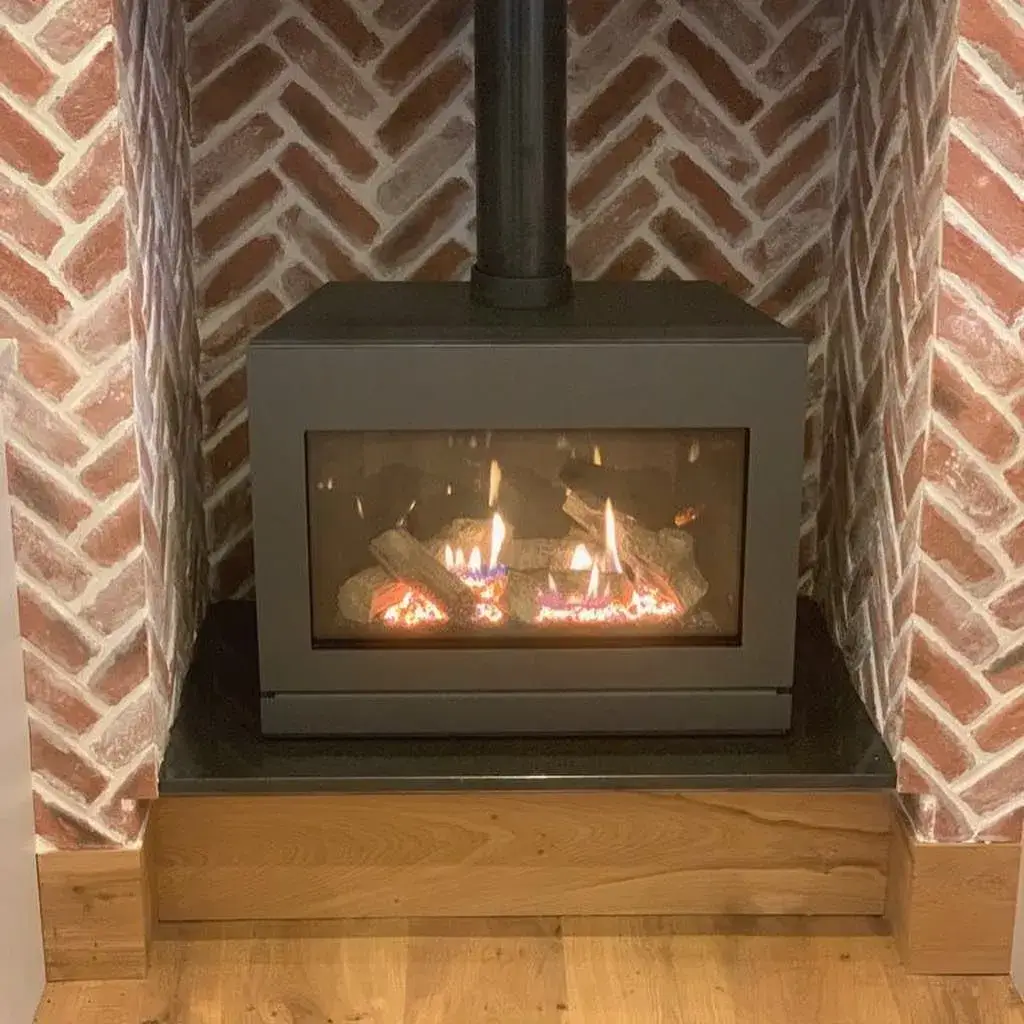 Black wood stove with visible flames inside, set against a red brick wall backdrop. The stove sits on a black base framed by a wooden border. The flooring in the room is light wood.