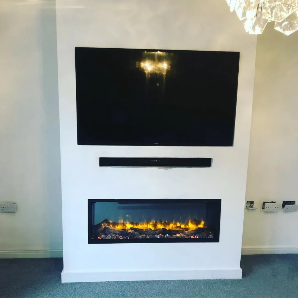 A modern living room setup featuring a mounted flat-screen TV above a slim black soundbar. Below is a rectangular electric fireplace with glowing embers and flames. The wall is white, and the flooring is a light gray carpet.