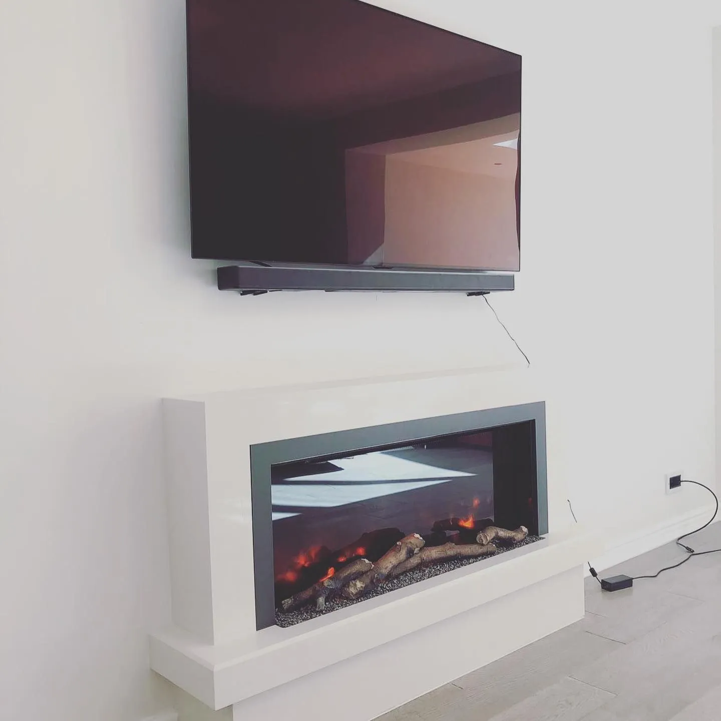 A modern living room with a flat-screen TV mounted on a white wall above a sleek, contemporary electric fireplace. The fireplace features realistic-looking logs and glowing embers. The setup is minimalistic and elegant.