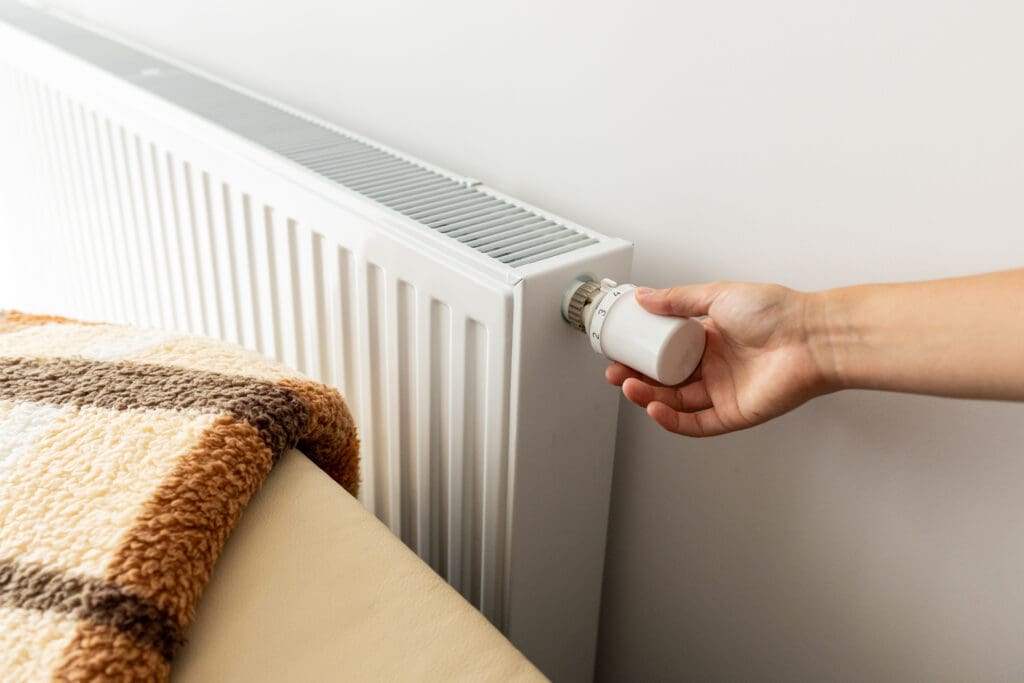 A hand adjusts the thermostat on a white radiator next to a chair with a striped blanket draped over it. The scene suggests a cozy indoor setting.