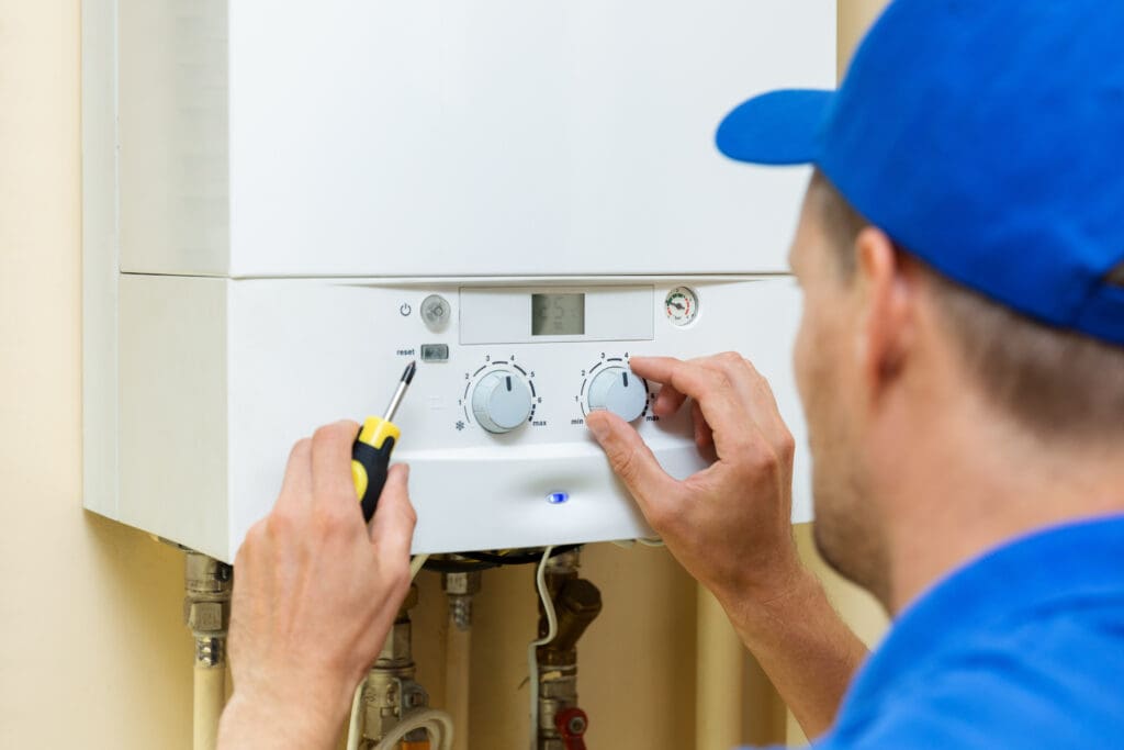 A person in a blue cap adjusts the settings on a white wall-mounted boiler using a screwdriver. The boiler's control panel displays dials and a small screen. Pipes are visible underneath the unit.