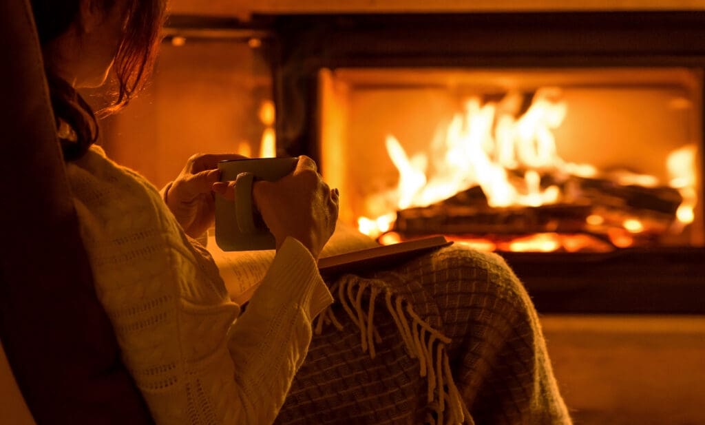 A person in a cozy sweater sits in front of a fireplace, holding a mug and wrapped in a blanket. The warm glow of the fire illuminates the scene, creating a comfortable and inviting atmosphere.