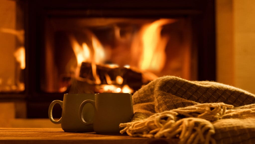 Two mugs sit on a wooden surface in front of a cozy, crackling fireplace. A warm, checkered blanket is draped beside them. The scene emits a warm, inviting glow, suggesting a peaceful, relaxing atmosphere.