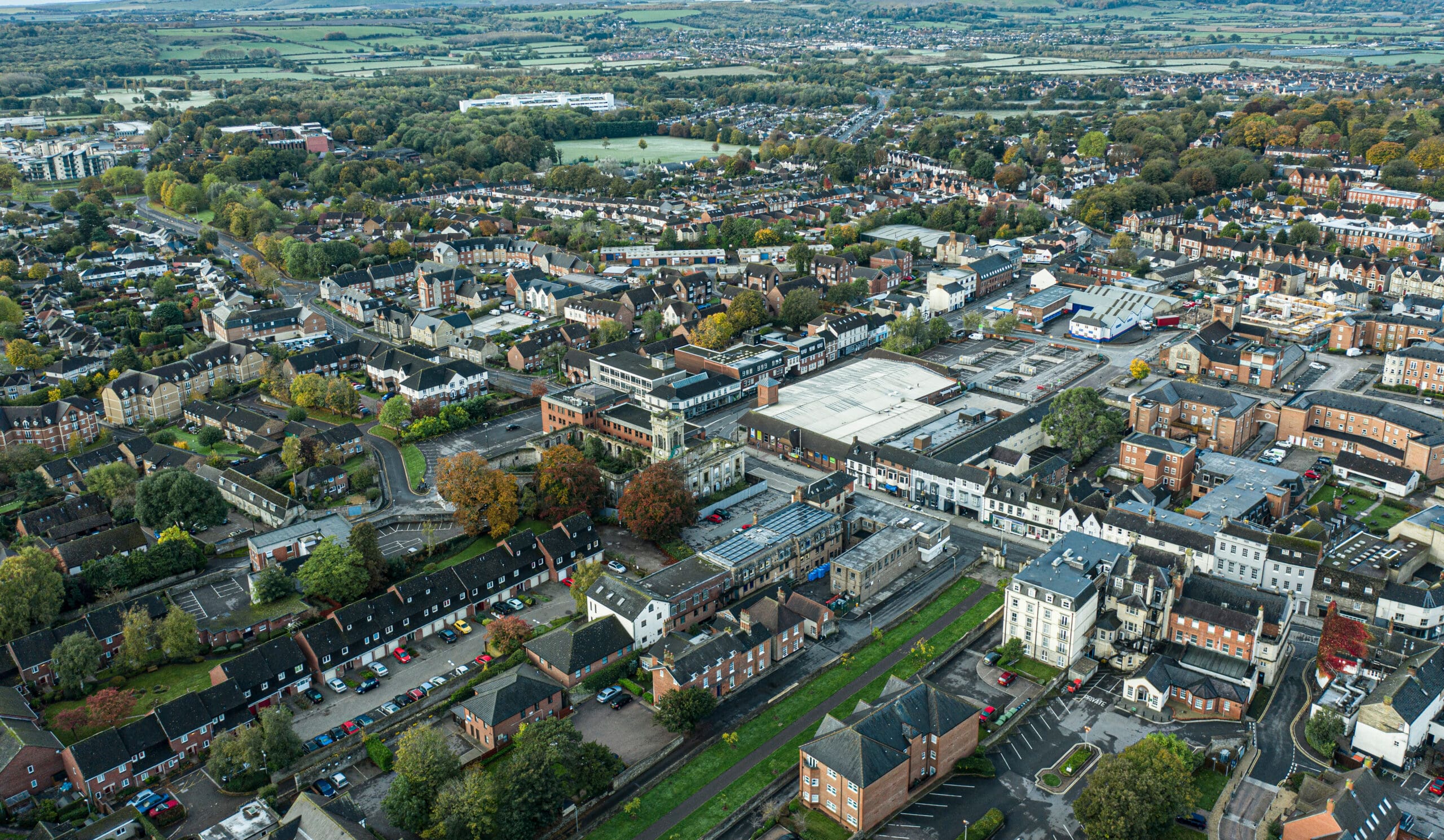 Aerial view of a town with diverse architecture, including residential and commercial buildings. Streets are lined with trees, and parking lots are visible. The landscape stretches into the distance with patches of greenery and open spaces.