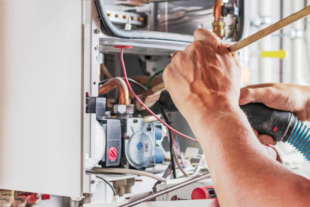 Close-up of a person working on a boiler with various tools. Visible are hands adjusting pipes and wires inside the open panel. Copper pipes and mechanical components can be seen, along with red and black wiring.