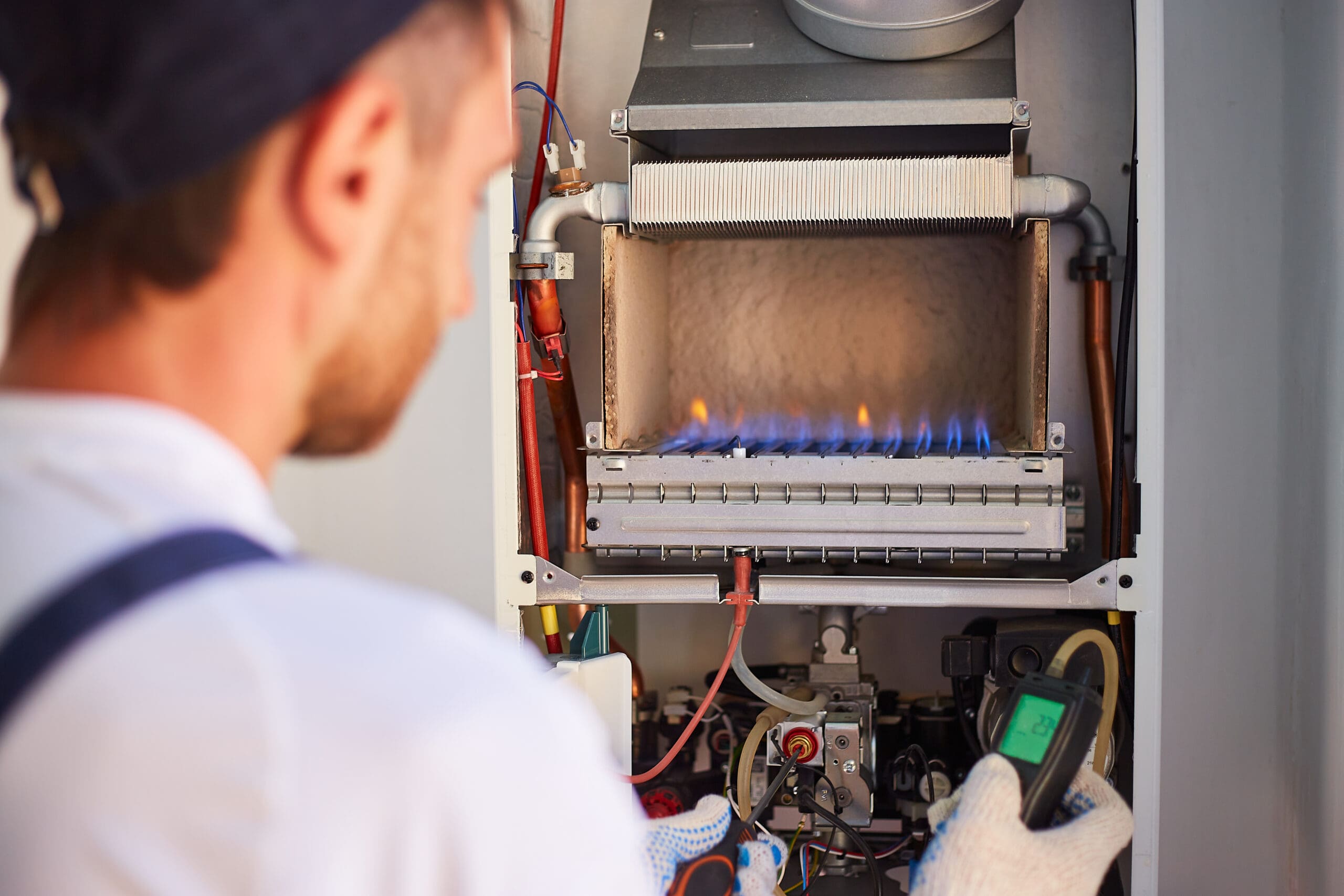 A technician in a cap and gloves is inspecting a gas furnace. The furnace's flames are visible, and he is using a handheld device to check its operation. Various pipes and components are exposed inside the open furnace panel.