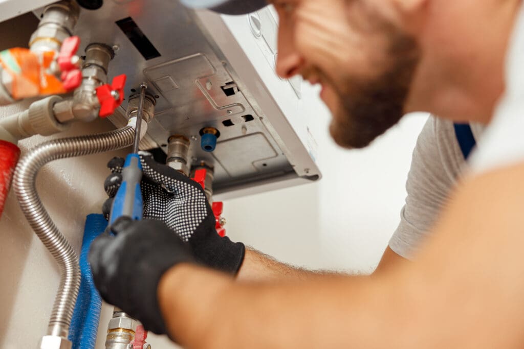 A plumber, wearing a cap and gloves, uses a wrench to adjust pipes connected to a metal panel. Various valves and fittings are visible, with red handles. The image focuses on the worker's hands and the plumbing components.