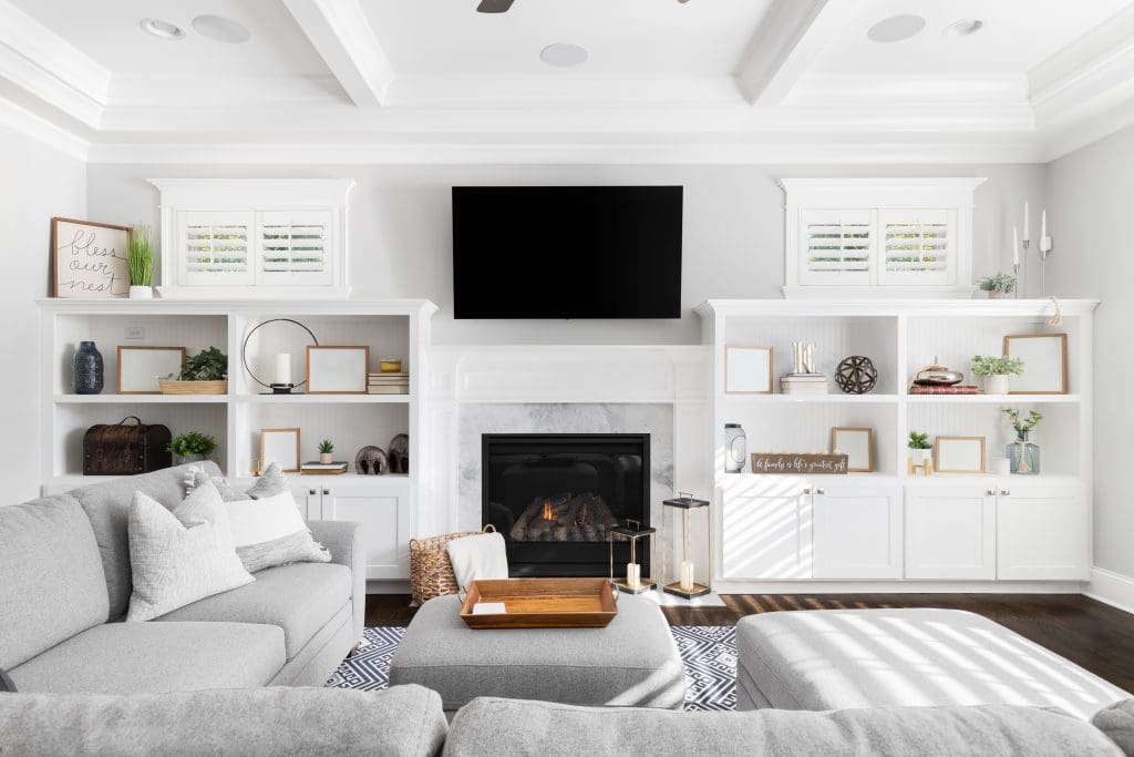 A modern living room with a light gray sectional sofa facing a fireplace. Above the fireplace is a large flat-screen TV. Built-in shelves on either side display books, plants, and decor items. The room features white walls and a coffered ceiling.