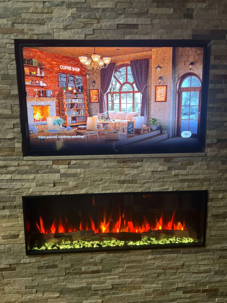 A television mounted above an electric fireplace displays a cozy coffee shop scene with warm lighting, bookshelves, and a seating area. The fireplace below features glowing red and yellow flames, adding warmth to the stone wall.