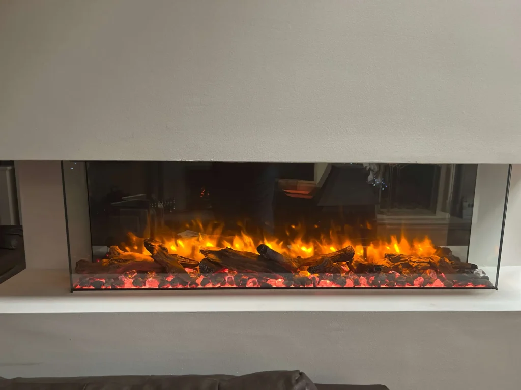 A modern electric fireplace with a wide glass front displays realistic orange flames and glowing ember logs. The fireplace is set within a neutral-colored wall, and a portion of a dark sofa is visible in the foreground.