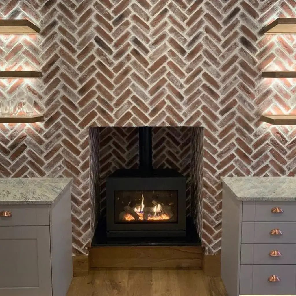 Modern fireplace with a brick herringbone pattern wall, surrounded by two shelves with soft lighting. The fireplace features a glass front showing a warm fire. Flanking the fireplace are two gray cabinets with copper handles, all on a wooden floor.