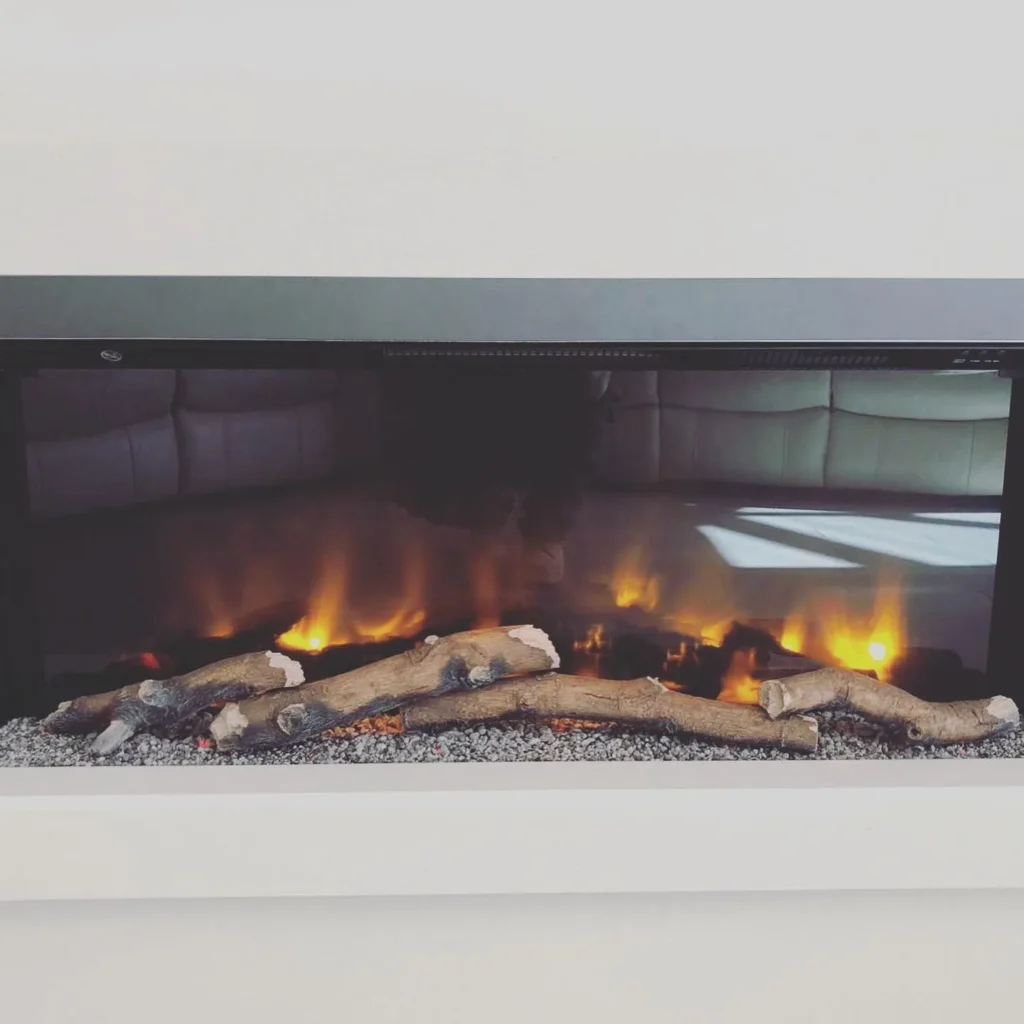 A modern electric fireplace set into a white wall. Artificial logs are placed on a bed of pebbles, with realistic flames flickering behind glass. A reflection of a white sofa is visible on the glass surface.