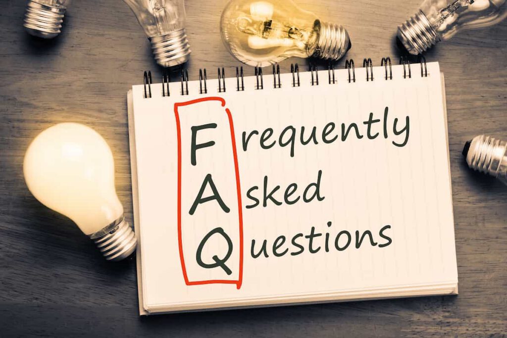 A notebook with "FAQ frequently asked questions" written on it, surrounded by lit and unlit lightbulbs on a wooden surface. The letters "FAQ" are highlighted in a red box.