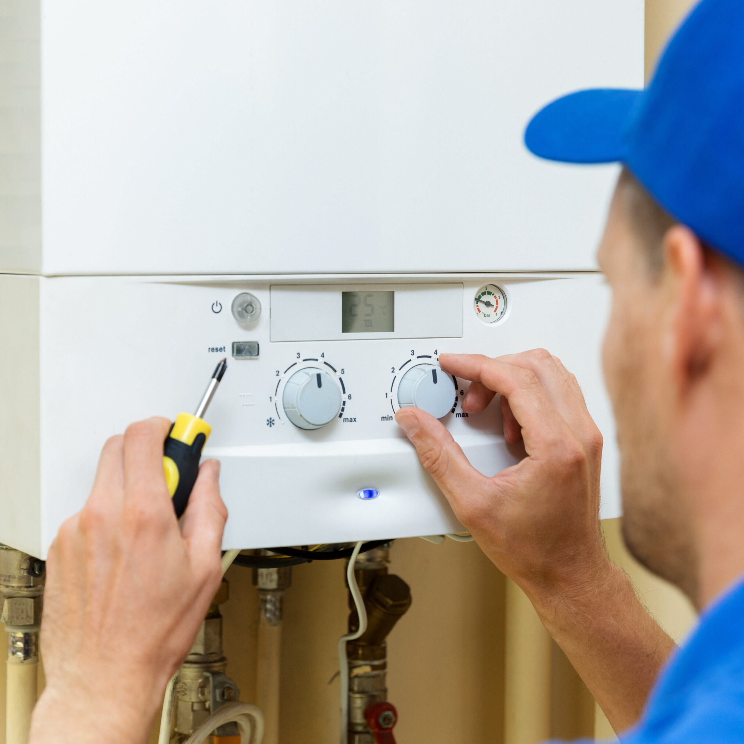 A person in a blue cap adjusts the settings on a white wall-mounted boiler using a screwdriver. The boiler's control panel displays dials and a small screen. Pipes are visible underneath the unit.