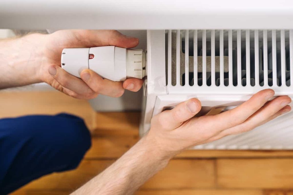 A person’s hands installing or adjusting a thermostat valve on a white radiator, with wooden flooring visible in the background.