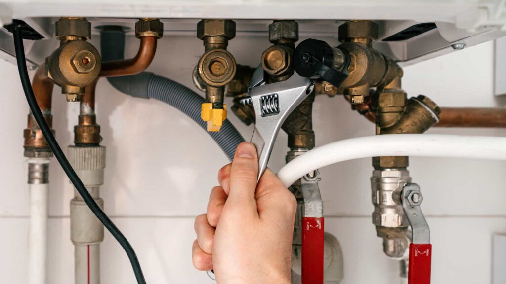 A hand uses a wrench to adjust a valve among several metal pipes and fittings, likely part of a plumbing or heating system.