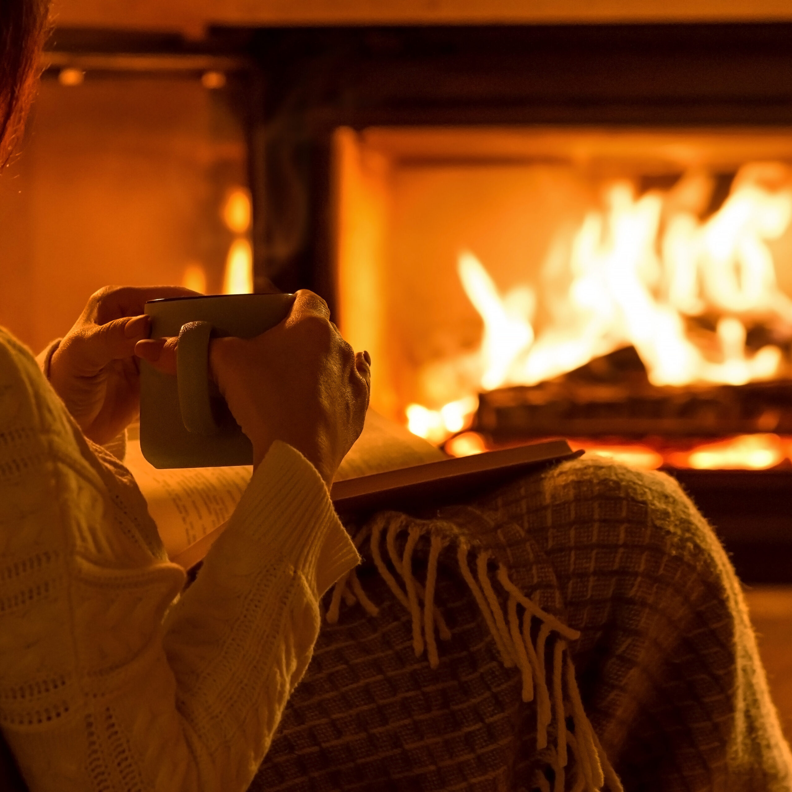 A person in a cozy sweater sits in front of a fireplace, holding a mug and wrapped in a blanket. The warm glow of the fire illuminates the scene, creating a comfortable and inviting atmosphere.