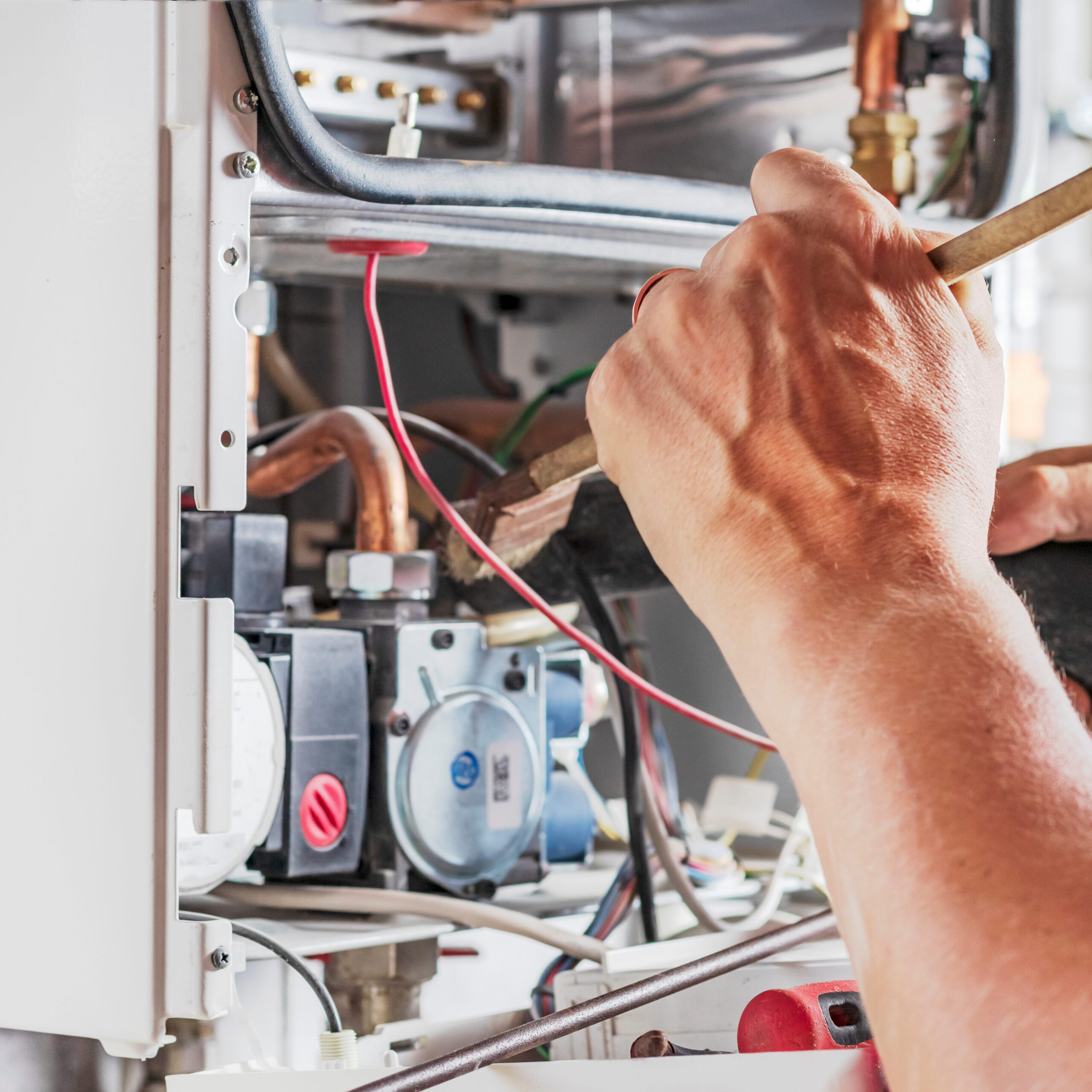 Close-up of a person working on a boiler with various tools. Visible are hands adjusting pipes and wires inside the open panel. Copper pipes and mechanical components can be seen, along with red and black wiring.