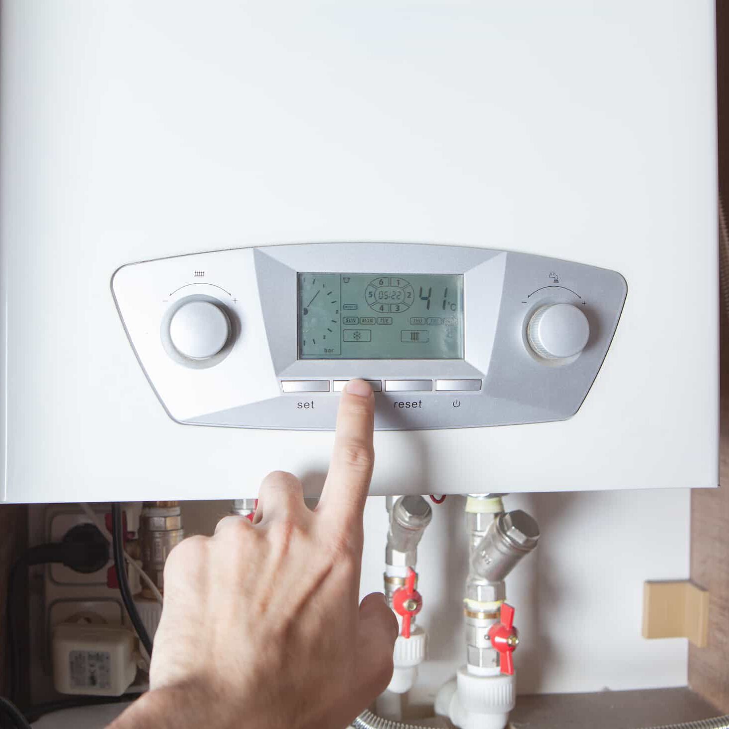 A person’s hand is pressing a button on the digital display panel of a modern boiler, with pipes and valves visible below the unit.