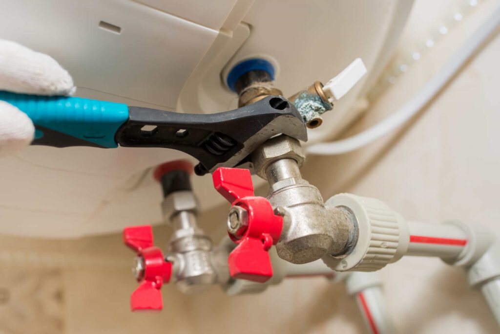 A gloved hand uses a wrench to tighten a metal valve connected to pipes with red handles, part of a plumbing or heating system.