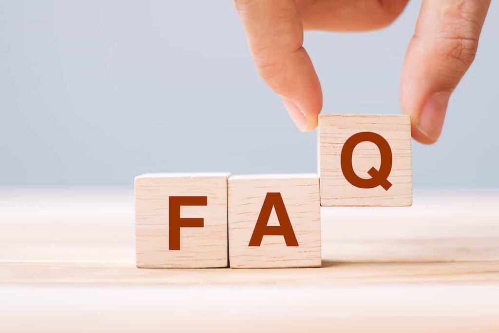 Three wooden blocks on a table spell out "FAQ." A hand is picking up the block with the letter "Q." The background is plain and softly lit.