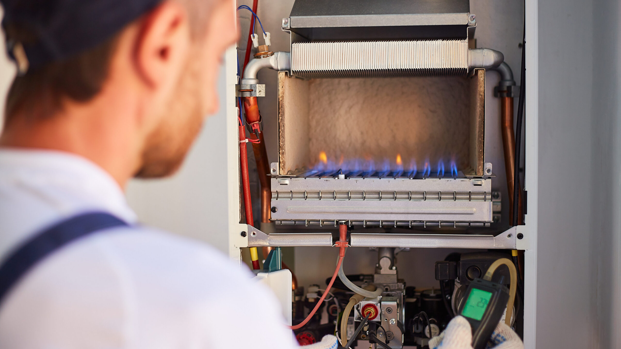 A technician in a cap and gloves is inspecting a gas furnace. The furnace's flames are visible, and he is using a handheld device to check its operation. Various pipes and components are exposed inside the open furnace panel.