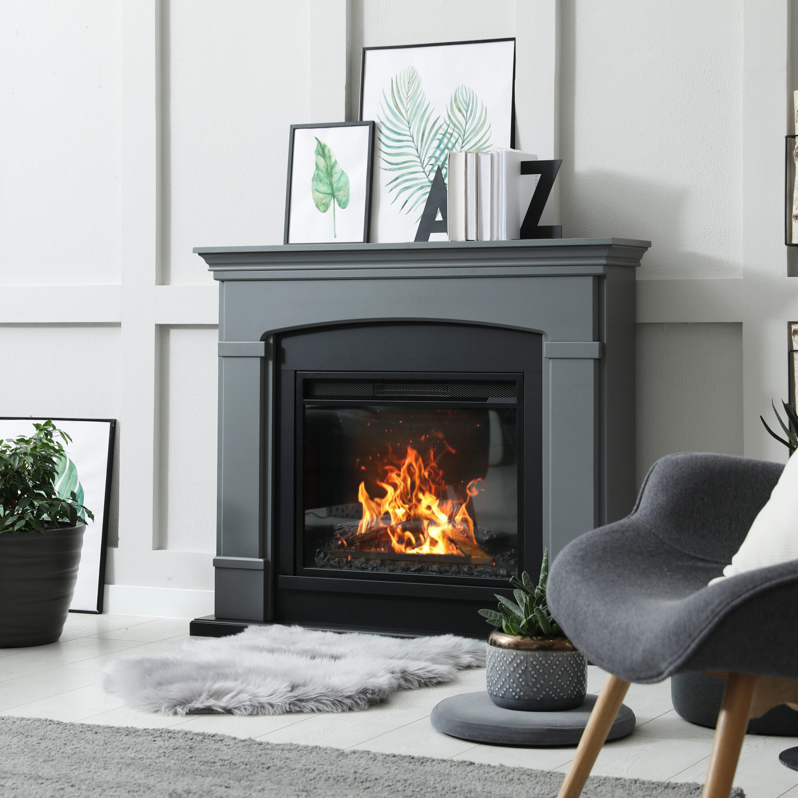 Modern living room with a sleek black fireplace and a burning fire. Grey chair and plush rug in front. Framed art and plants decorate the space, with a stack of firewood beside the mantel. Light-colored walls and floor provide a cozy ambiance.