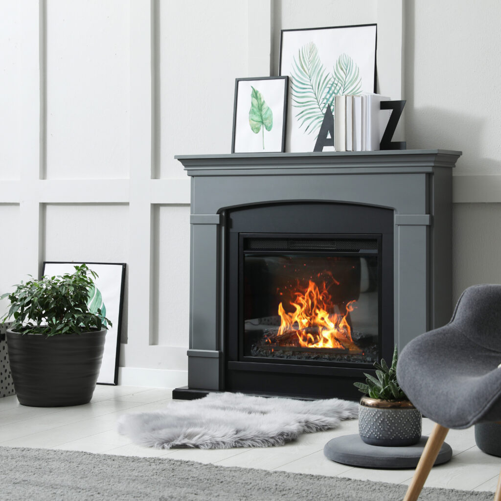 Modern living room with a sleek black fireplace and a burning fire. Grey chair and plush rug in front. Framed art and plants decorate the space, with a stack of firewood beside the mantel. Light-colored walls and floor provide a cozy ambiance.