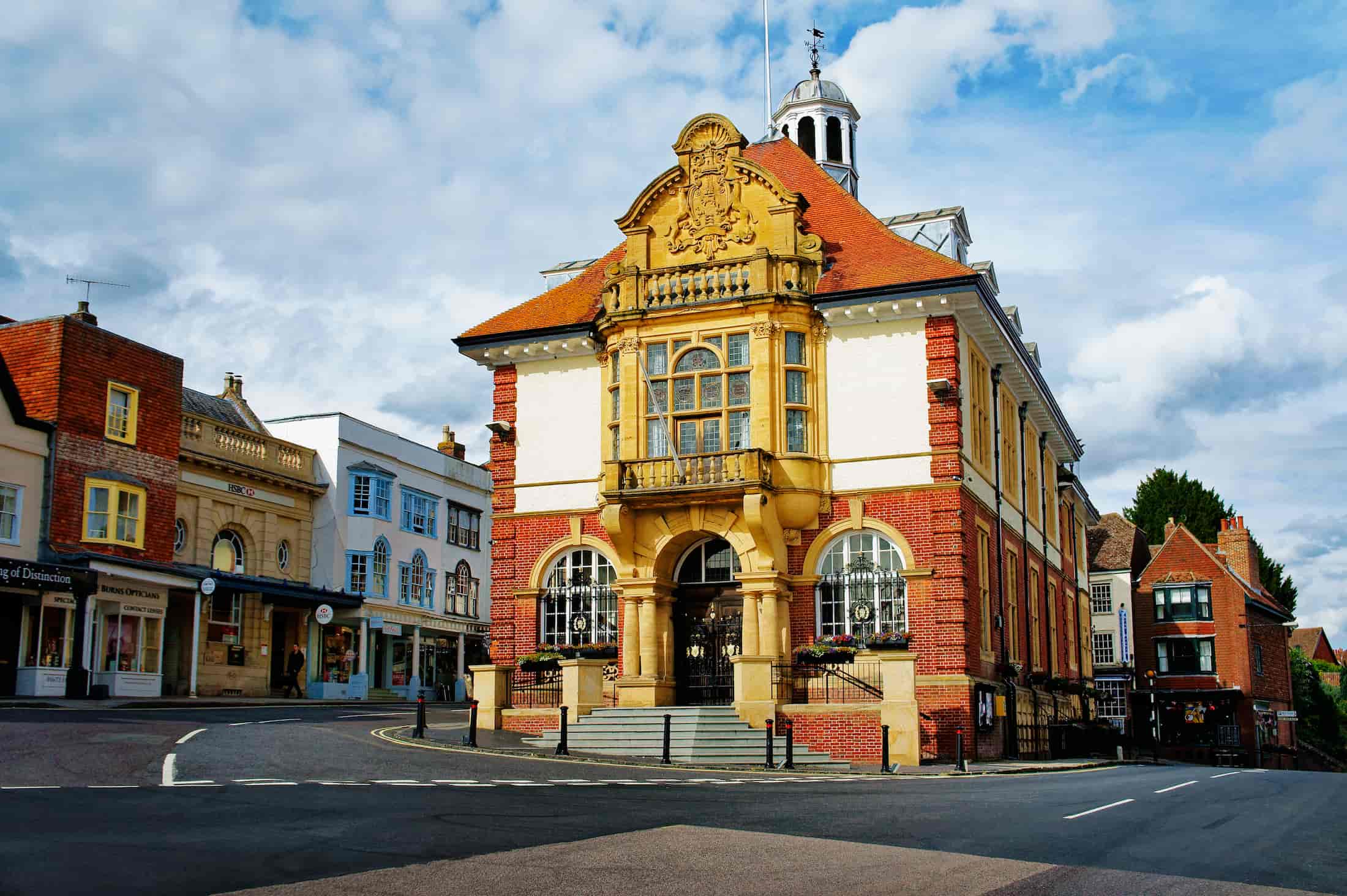 A historic building with ornate architecture and a red-tiled roof stands at a street corner, surrounded by smaller shops and houses under a partly cloudy sky.