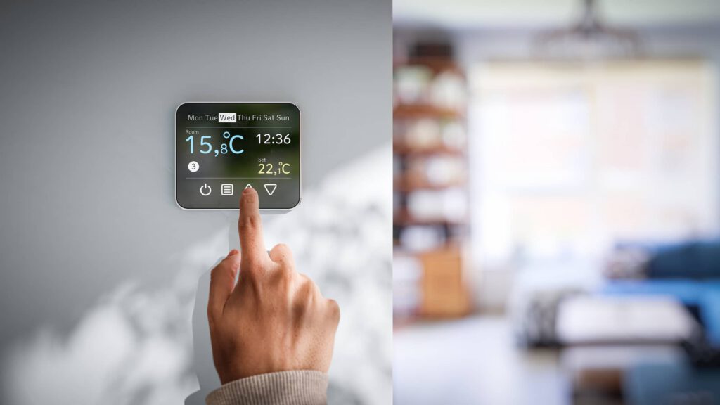 A hand presses a button on a digital smart thermostat mounted on a wall, displaying the date, time, and indoor and outdoor temperatures, with a blurred modern living room in the background.