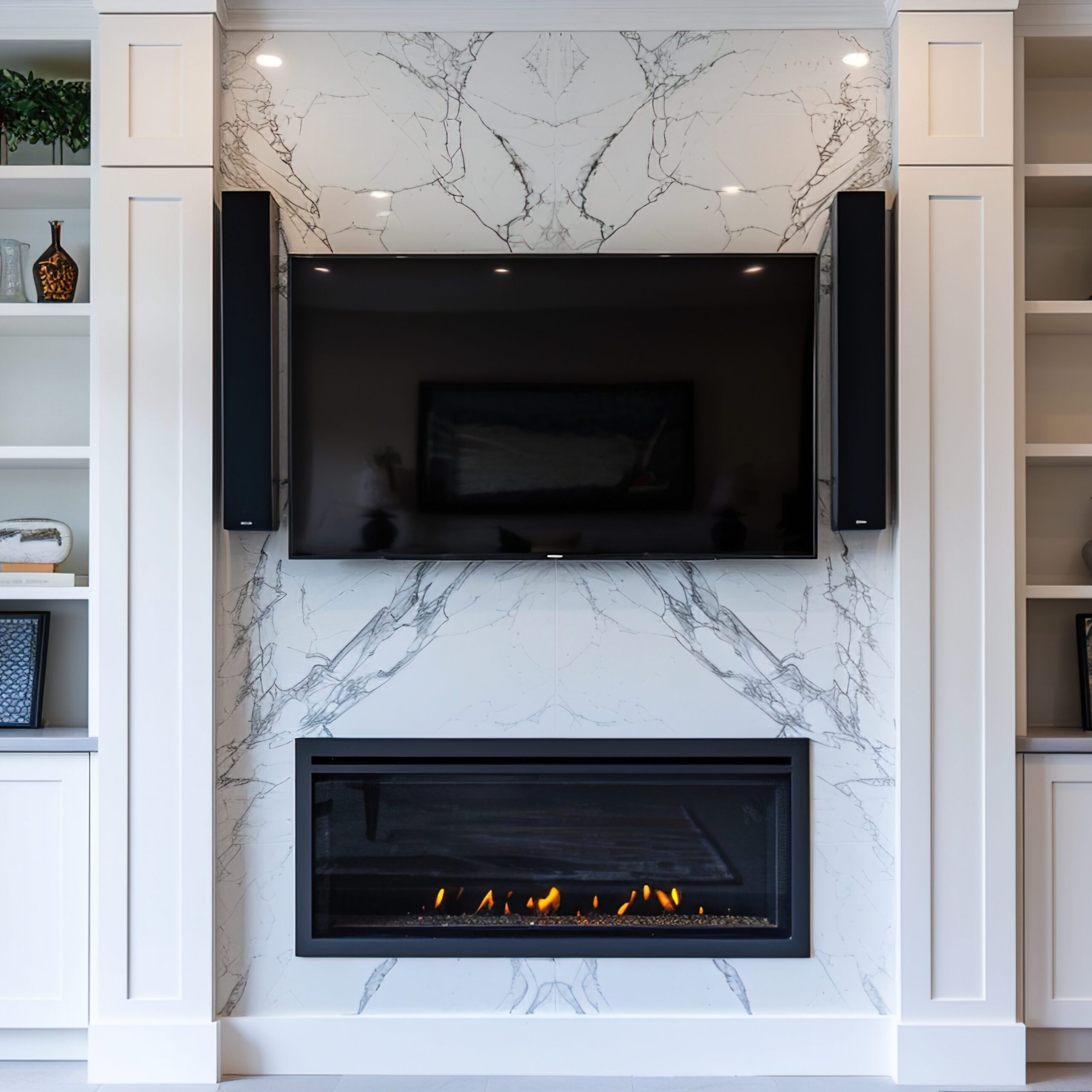 Modern living room with a wall-mounted TV above a sleek electric fireplace set in a marble facade. White built-in shelves and cabinets surround the area, displaying decorative objects and plants. Cozy gray sofas flank the fireplace.
