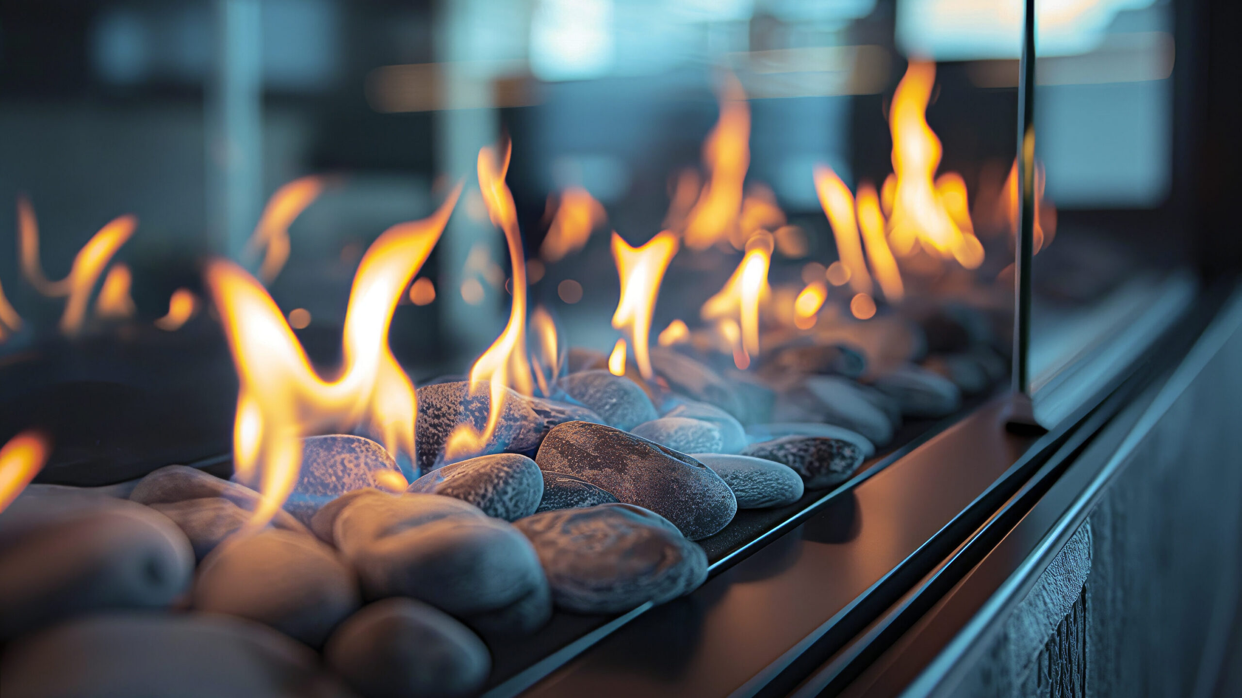 Decorative fire flames dance over smooth gray stones in a modern glass-enclosed fireplace, creating a warm and cozy ambiance. The background is softly blurred, highlighting the vibrant orange and yellow hues of the flames.